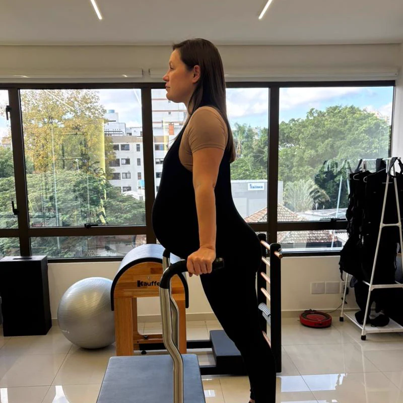 Gestante fazendo Pilates em pé no aparelho Chair, com as mãos na barra